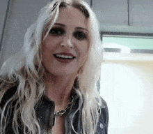 a blonde woman wearing a leather jacket and a necklace smiles