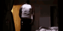 a man in a white tank top is standing in a dark room next to a door .