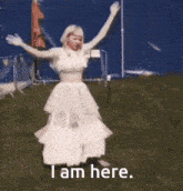 a woman in a white dress is standing in the grass with her arms outstretched and the words i am here .