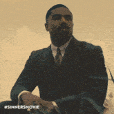 a man in a suit and tie sits in the grass with #sinnersmovie written on the bottom