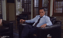 a man in a blue shirt and tie is sitting in a chair with a glass of whiskey in his hand ..