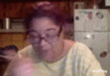 a woman wearing glasses and a yellow shirt is sitting in front of a refrigerator in a kitchen .