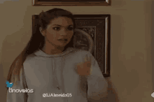a woman in a white hoodie is giving a thumbs up while standing in front of a wall .