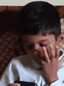 a young boy is covering his face with his hand while looking at his cell phone .