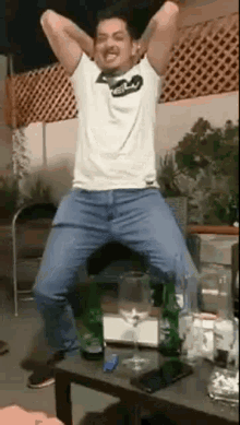 a man in a white shirt and blue jeans is dancing in front of a table with bottles of alcohol on it .