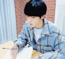 a young man in a plaid shirt is sitting at a table with a pen in his hand