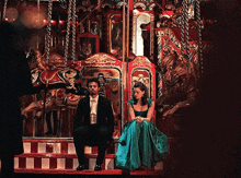 a man in a suit sits next to a woman in a blue dress in front of a merry go round