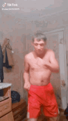 a shirtless man in red shorts is dancing in a room with a dresser .