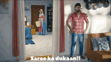 a man in a plaid shirt is standing in a living room with the words saree ka dukaan on the bottom right