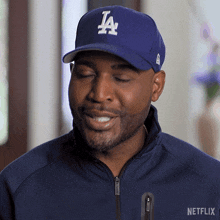 a man wearing a la hat and a netflix jacket smiles with his eyes closed