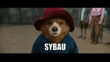 a paddington bear is wearing a red hat and a blue shirt and says sybau .
