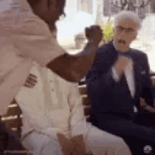 a man in a suit is talking to another man in a white shirt while sitting on a bench .