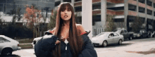 ariana grande is wearing a black jacket and standing in a parking lot with cars .