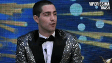 a man in a tuxedo and bow tie is talking into a microphone with the words viperissima trash behind him