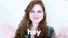 a woman with long red hair is smiling and waving at the camera while saying hey .