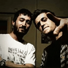 two men are standing next to each other in a dark room . one of the men is pointing at the camera .