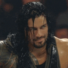 roman reigns is a wrestler with long hair and a beard and is looking at the camera .