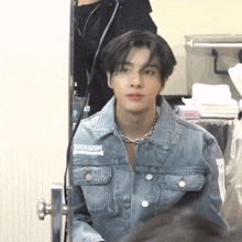 a man wearing a denim jacket and a necklace is looking at himself in a mirror .
