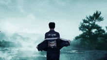 a man wearing a jacket that says jh jung stands in front of a river