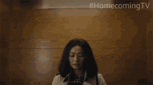 a woman in a trench coat stands in front of a wooden wall with #homecoming tv written on the bottom