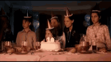 a group of children wearing party hats are sitting around a table with a birthday cake .