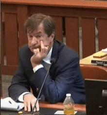 a man in a suit sits at a desk with a microphone and a bottle of water