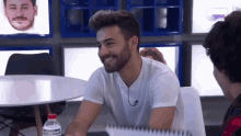 a man with a beard is smiling while sitting at a table with a notebook and a water bottle .