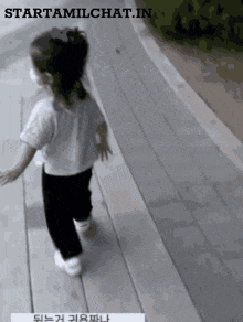 a little girl wearing a white shirt and black pants is walking down a wooden walkway