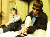 a man wearing sunglasses is sitting on a bed next to another man