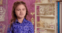 a young girl in a blue shirt is making a funny face in front of a shelf full of crowns .