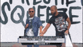 two men holding hands in front of a wall that says squeeze