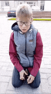 a boy wearing glasses and a vest is kneeling down on the ground .