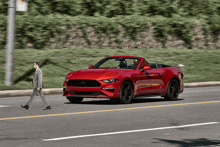 a man crosses the street in front of a red convertible ford mustang