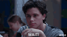 a young man is sitting at a table with a pencil in his hand and the words wait written on his face