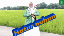 a woman in a green jacket stands in front of a field of green grass and a sign that says ' atur nuhun '