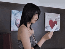 a woman holds a piece of paper in front of two paintings one of which has a heart on it