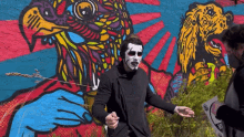 a man with white face paint stands in front of a colorful wall mural