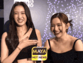 two women standing next to each other with a maya hot news sign in front of them