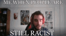 a man wearing headphones is sitting in front of a wall with posters on it and says me when people are still racist