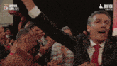 a man in a suit and tie stands in front of a crowd with his arms in the air in front of a benfica logo