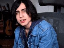 a young man wearing a denim jacket and plaid shirt is sitting in a chair with a guitar in the background