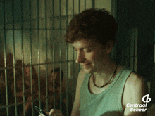 a man in a green tank top is looking at a cell phone with a central beheer logo in the corner