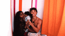 a man without a shirt is holding a baby and a woman kisses him on the cheek