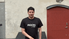 a man wearing a black shirt that says agon by aoc stands in front of a red door