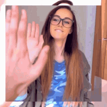 a woman wearing glasses and a blue camo shirt waves her hand