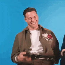 a man in a green jacket is laughing with his eyes closed while holding a remote control .