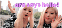 two blonde women are waving their hands in front of a sign that says ara says hello !