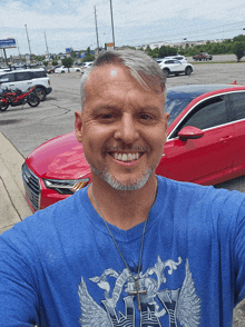 a man wearing a blue shirt with the word why on it smiles in front of a red car