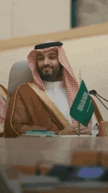 a man in a saudi arabian robe is sitting at a table holding a green flag