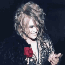 a man with long curly hair is holding a red rose in his hand and smiling .